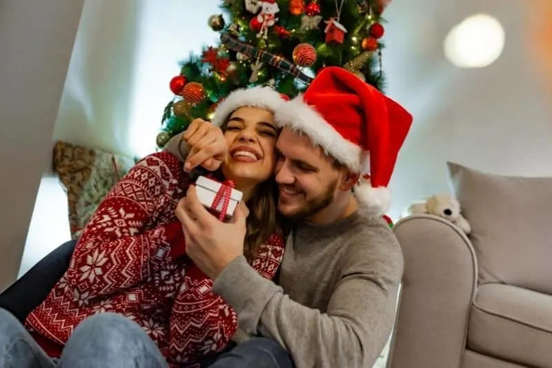 sweet couple opening gift inside home with a christmas tree behind them