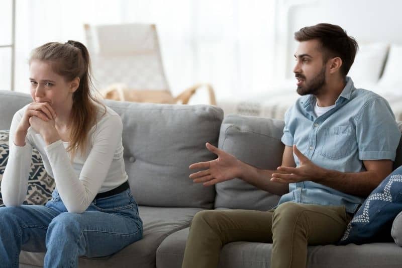 upset man lecturing a crying woman sitting on living room's couch