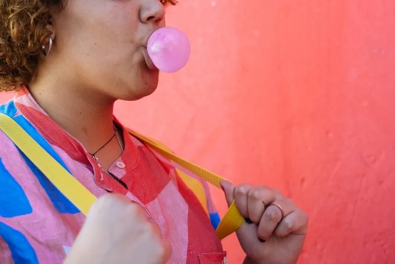 woman blowing gum bubble while standing near pink wall