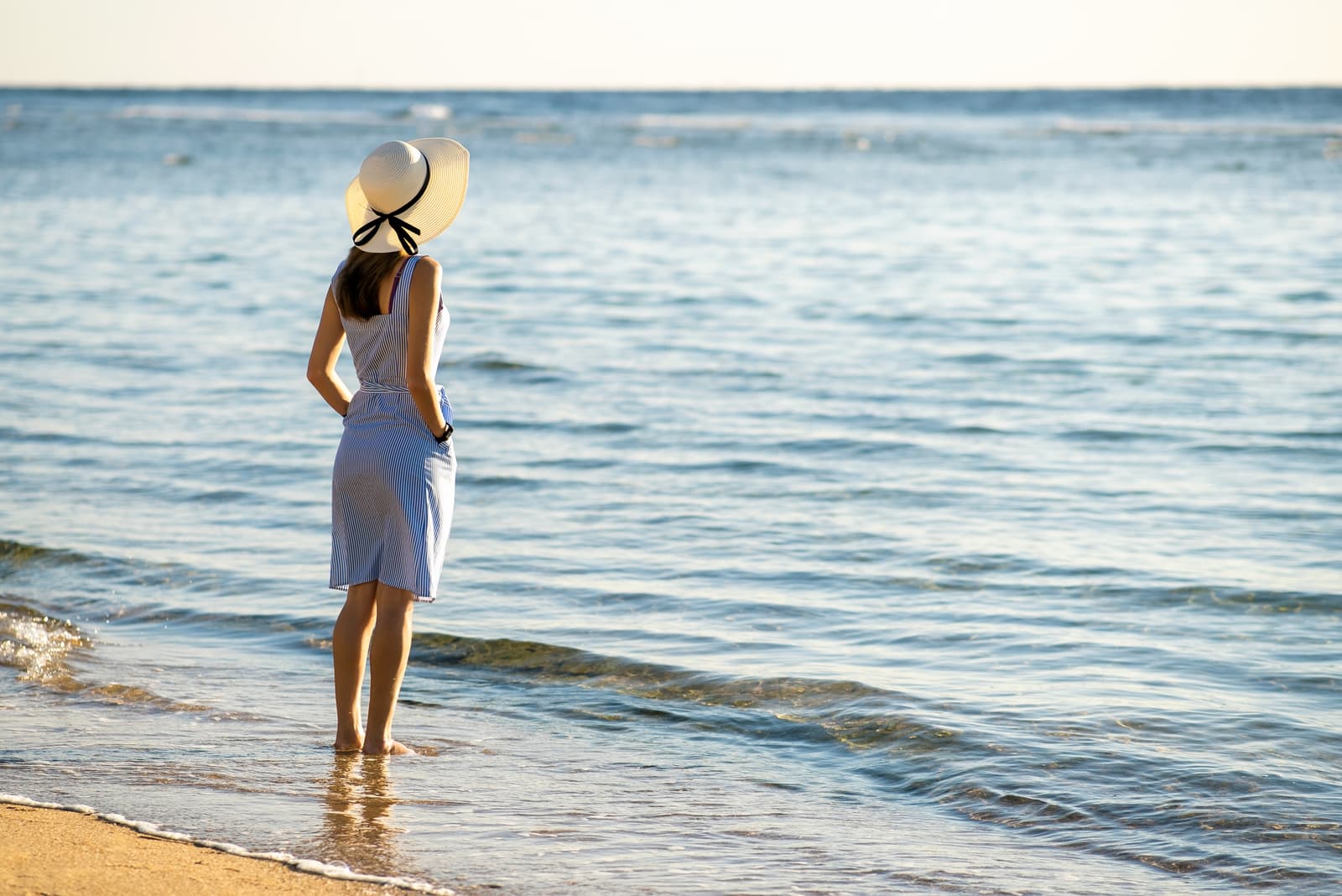  woman in straw hat and a dress standing alone on empty sand beach