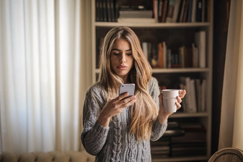 woman looking at phone while holding white mug