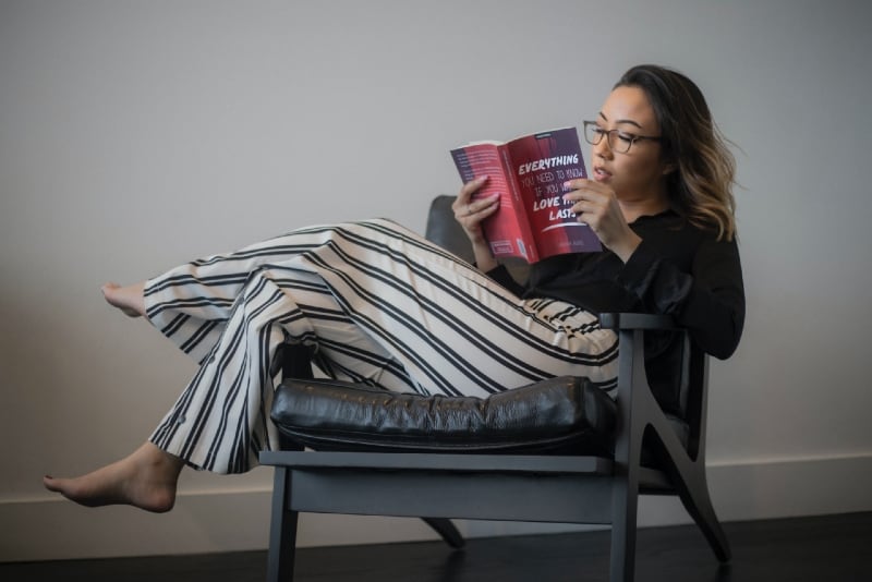 woman with eyeglasses reading book while sitting on chair