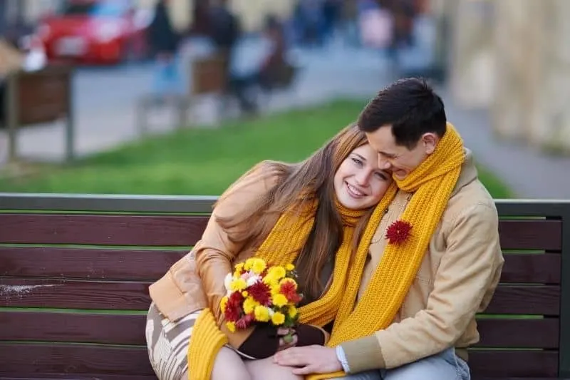 woman receiving a bouquet of flower from her man sitting and hugging her on the bench outdoors