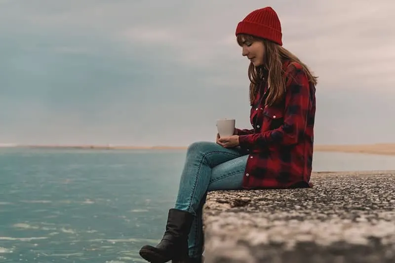 woman sitting near the sea holding a mug wearing red bonnet and checkered top