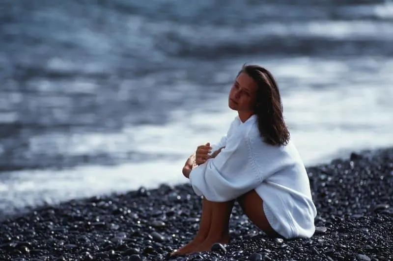 woman sitting pensively on the small rocks on the seashore wearing white big top