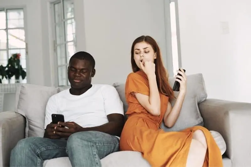 woman sneaking on man's phone while sitting on couch and holding her phone inside home