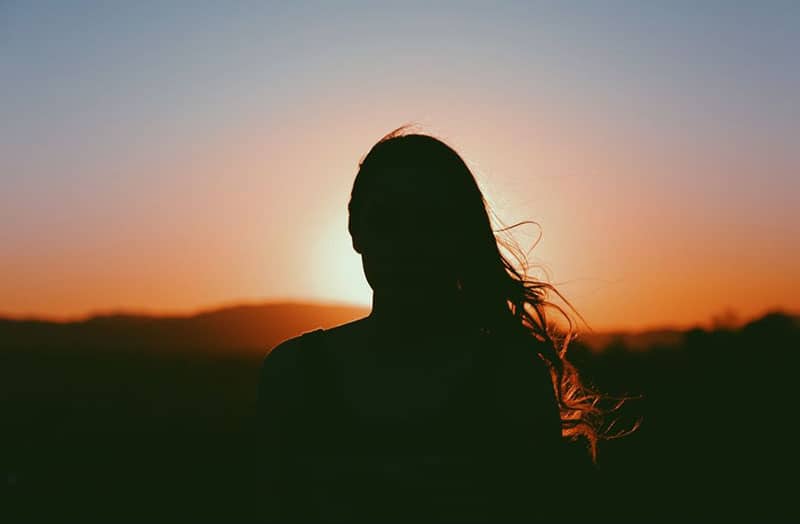 Immagine di donna al tramonto in piedi e rivolta verso il sole che tramonta con i suoi raggi arancioni