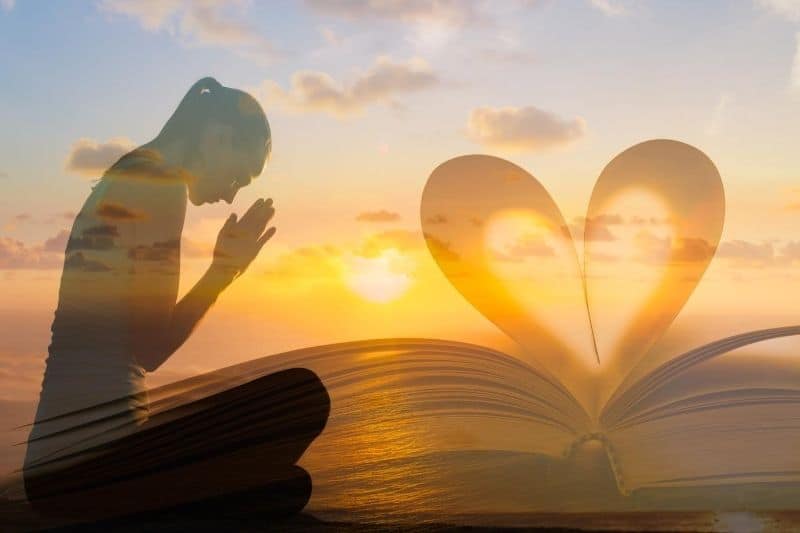joven mujer rezando junto a una biblia en forma de corazón contra la puesta de sol