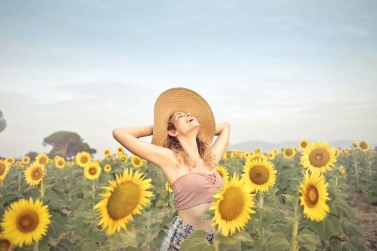 mulher sorridente com chapéu, de pé num campo de girassóis