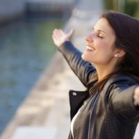 grateful woman wearing black jacket raising arms standing on river bank