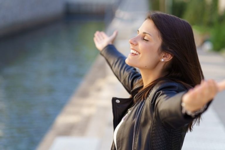 grateful woman wearing black jacket raising arms standing on river bank
