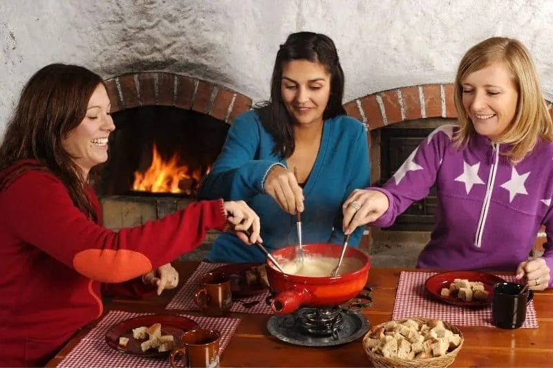3 beautiful ladies dipping bread in fondue inside home near a fireplace