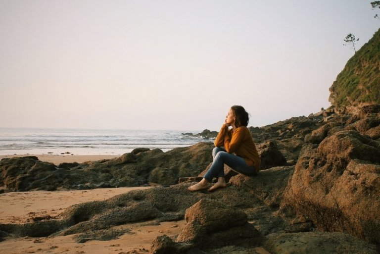 woman in yellow sweater sitting on rock looking at sea