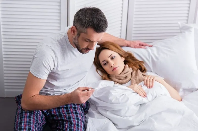 Boyfriend looking at the temperature in the thermometer with a sick woman beside him lying on bed