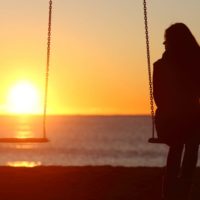 silueta de una mujer sentada en un columpio sola mirando un columpio vacío a su lado contra el amanecer cerca de la playa