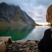 mujer con chaqueta negra sentada en una roca cerca de un lago