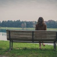 vista posterior de una mujer sentada sola en un banco frente a una masa de agua