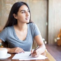 mulher a escrever num papel e a pensar enquanto olha para longe dentro de um café