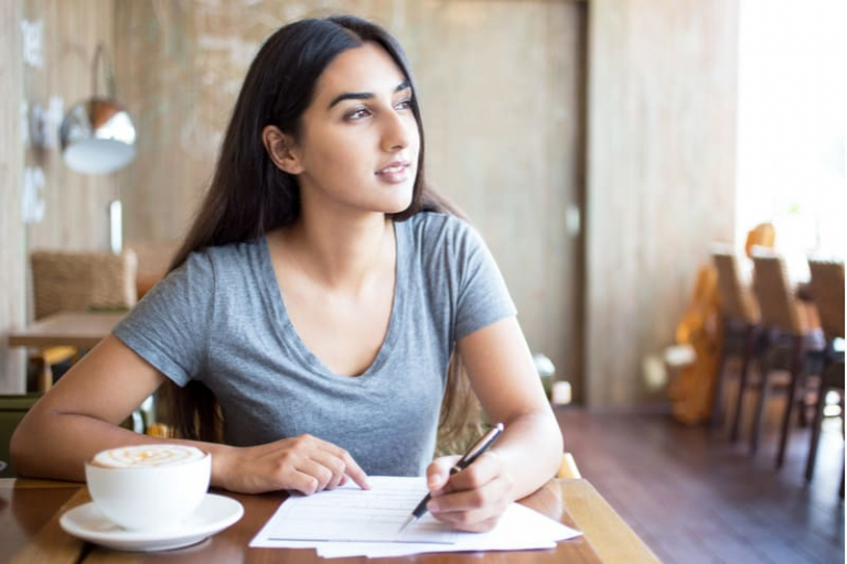 woman writing down on a paper thinking while looking far inside a cafe