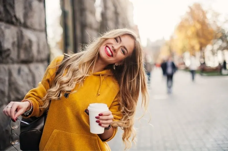 cheerful woman smiling bending her head slightly to the right walking outdoors with coffee on her hand