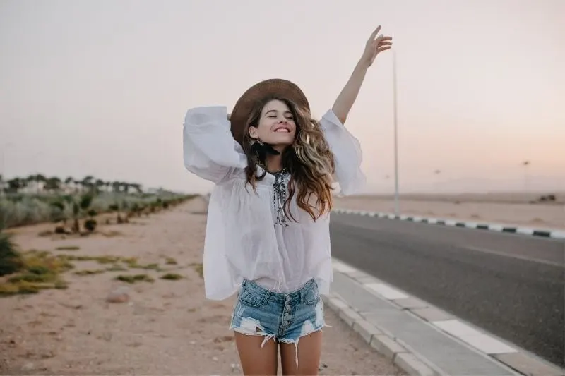 cheerful young woman raising her hand and holding hat standing beside the highway