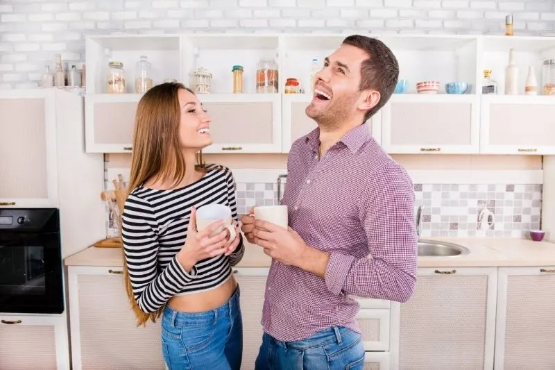 couple laughing hard inside the kitchen while holding coffee