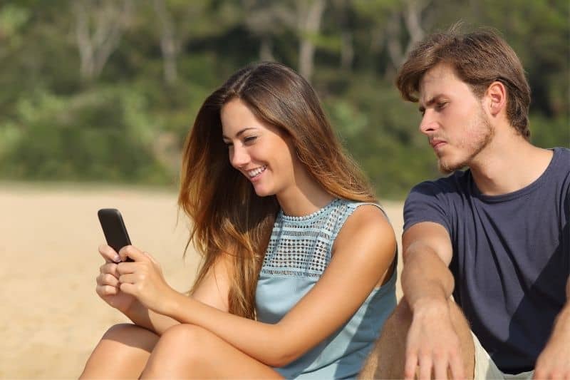 Fidanzato geloso che guarda i messaggi della sua ragazza mentre è seduto in spiaggia