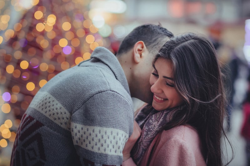 homem a beijar uma mulher ao nível do pescoço, ao ar livre, perto de uma grande árvore de Natal