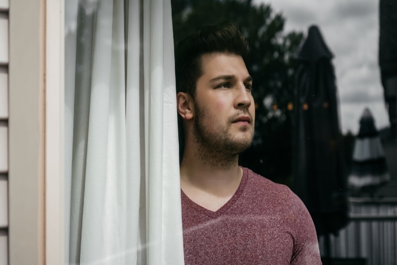 man looking through window while standing near white curtain