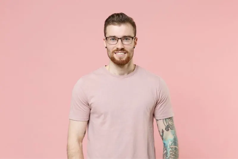 man with tattoo and ear piercing wearing eyeglasses and pink shirt standing against a pink wall