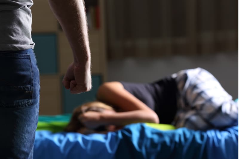 mistreated victim lying on bed with a cropped man standing with close fist