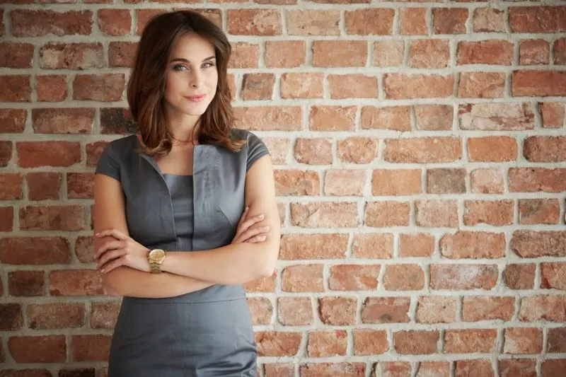 pensive woman standing against brown brick wall with arms crossed