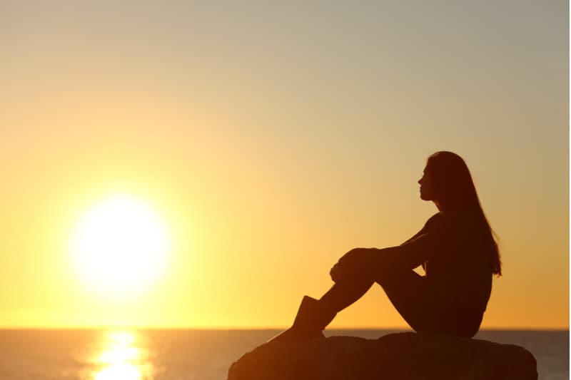 perfil de uma silhueta de mulher sentada numa rocha perto da água durante o pôr do sol