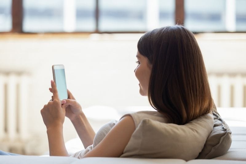 vista trasera de una mujer millennial feliz enviando mensajes por el móvil tumbada en el sofá