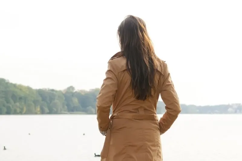 rear view of a woman standing in front of a body of water wearing brown trench coat