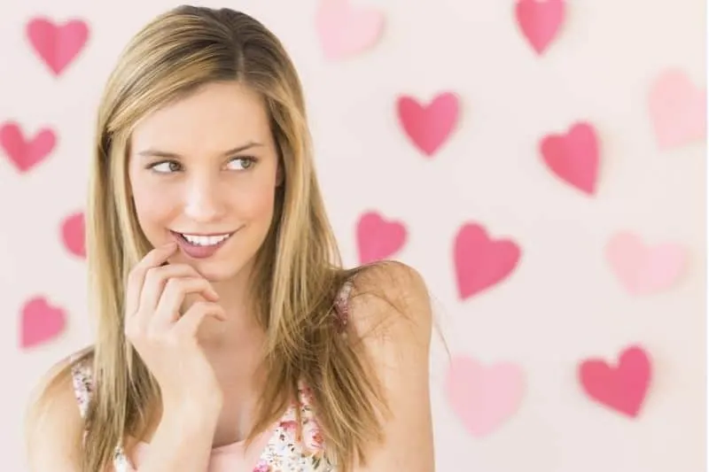 shy woman biting lips and smiling looking at her left standing against a wall with pink hearts