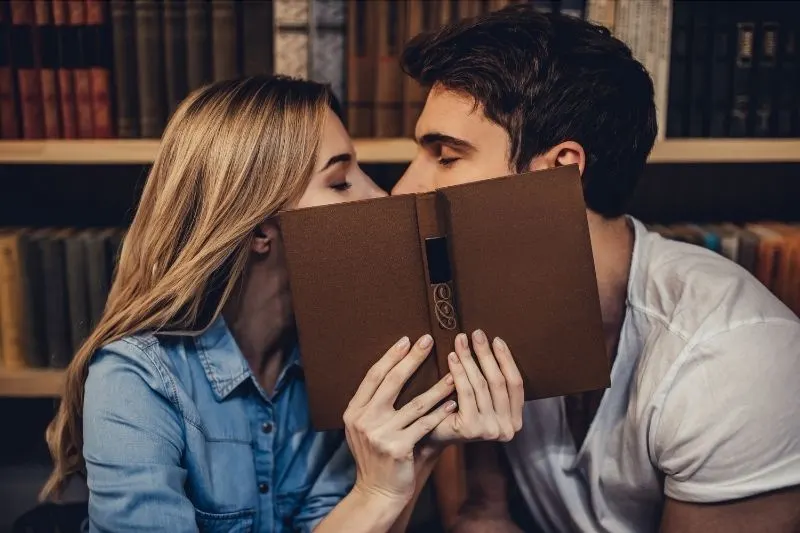 students kissing inside the library covering face with a book