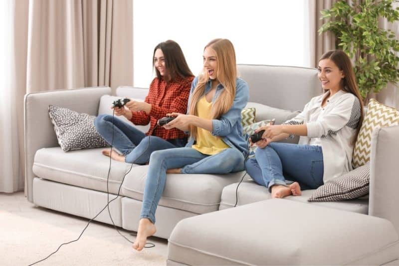 three friends playing video games inside home sitting on the sofa