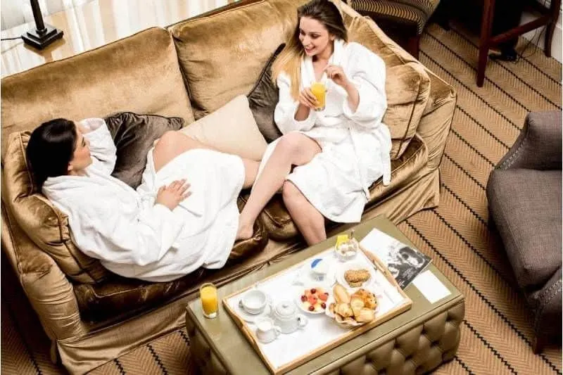 two friends chatting after a spa wearing white robes relaxing at the sofa and snacks on the table