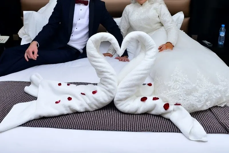 two towels swan shaped in hotel bed with a newly wed couple cropped at the back