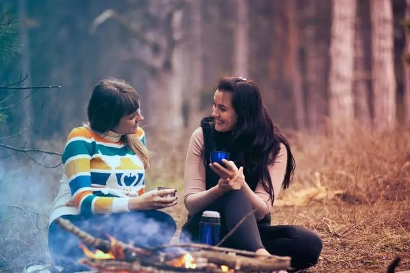two women sitting on the ground near a bonfire in the forest