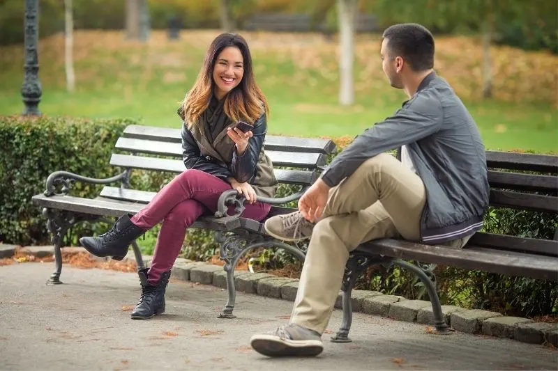 two young people sitting on a separate benches of the park talking and smiling