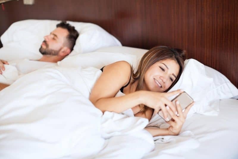 woman busy texting on bed beside her sleeping husband