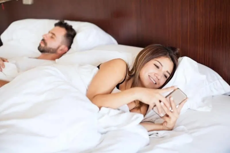 woman busy texting on bed beside her sleeping husband