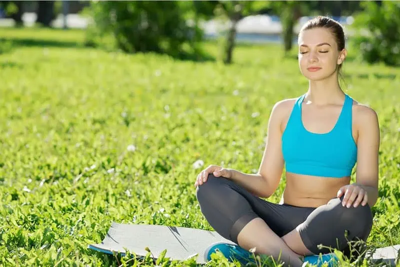 woman in yoga pose meditating outdoors with a mat on top of the green grasses