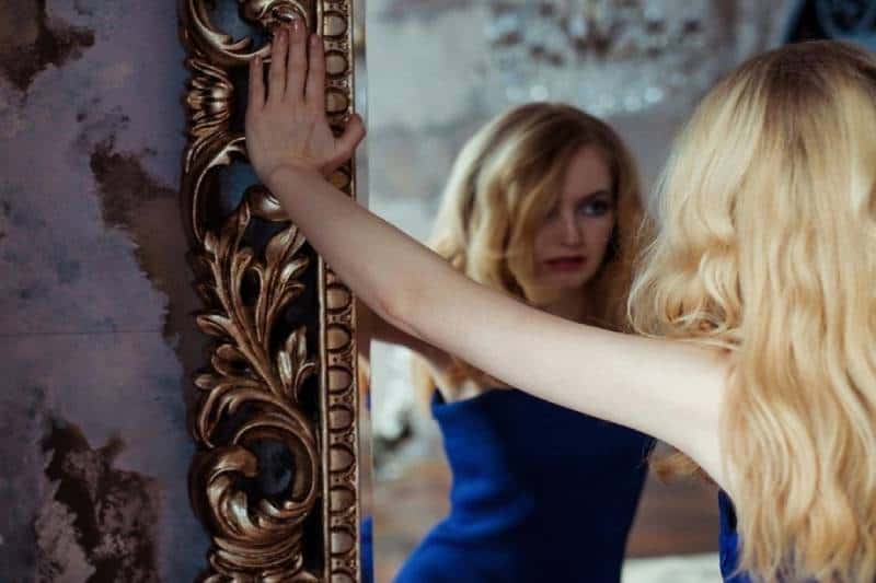 donna che si guarda allo specchio con indosso un vestito blu e le mani sul bordo dello specchio che pensa