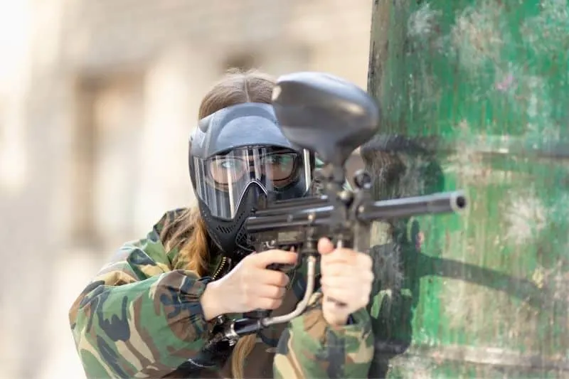 woman playing paintball with complete geared overall leaning and hiding ready to shoot