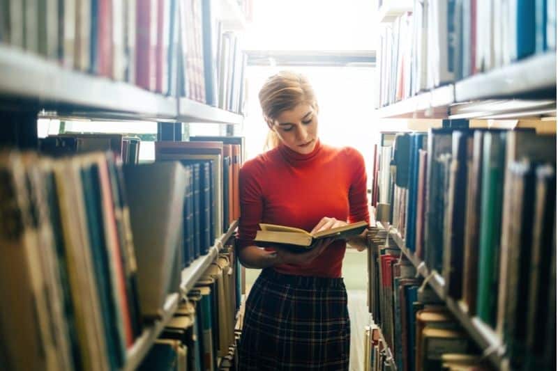 donna che legge in biblioteca in piedi in mezzo agli scaffali dei libri