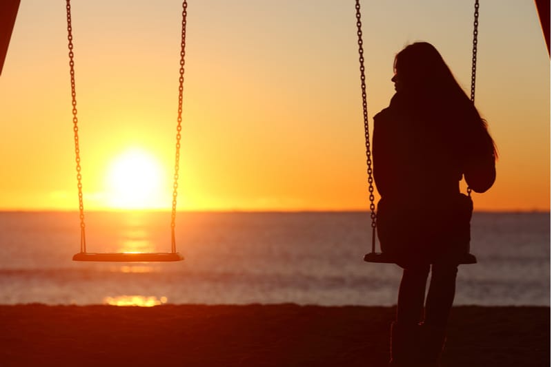 mujer sentada en un columpio con un columpio vacío detrás de ella frente al mar y la puesta de sol