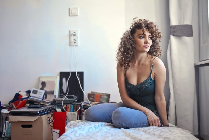 woman with curly hair sitting on bed looking through window
